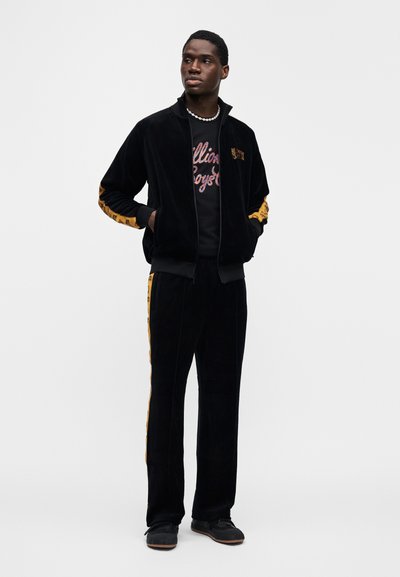 Μαύρο βελούδινο φόρμα με κίτρινες πλαϊνές λωρίδες, ζακέτα με φερμουάρ και αντίθετη μπλούζα με στρογγυλή λαιμόκοψη. Το μοντέλο φοράει μαύρα αθλητικά παπούτσια.