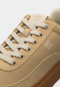 Μπεζ σουέντ αθλητικό παπούτσι με λευκά κορδόνια, καφέ σόλα και το λογότυπο "HUGO" στην πλευρά και τη γλώσσα.