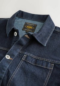 Denim shirt with a dark blue color, featuring a classic collar, metal buttons, and contrasting gold stitching. Label reads "Premium Grade 1982."