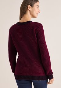 Knit svetr v tmavě burgundské barvě s texturovaným vzorem, žebrovaným výstřihem a manžetami a delším zadním spodním lemem, kombinovaný s námořnickými džíny.