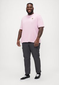 Tricou scurt cu mâneci scurte de culoare roz deschis, cu un mic logo galben, asortat cu pantaloni cargo de culoare gri închis și adidași negri. Potrivire casual, textură netedă.
