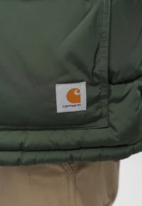 Roheline puhvjakker, millel on silmapaistev tikitud tekstuur, millel on valge ruudukujuline silt oranži logoga. Õmbluste detailid on nähtavad.