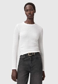 Witte textuur lange mouwen top met een ronde hals, gedragen met donkergrijze jeans en een zwarte riem met een gouden gesp.