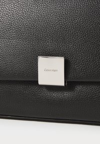 Crna kožna torba s teksturiranom površinom i pravokutnom srebrnom metalnom kopčom s brandom "Calvin Klein".