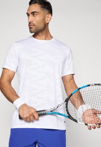 Mand i hvid mønstret skjorte og blå shorts holder en sort og blå tennisracket, iført hvide håndledsbånd, kigger til siden.