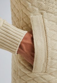Pleteni pulover u krem boji s teksturiranim dijamantnim uzorkom, s rebrastim rukavima i bočnom džepom. Mekan, udoban materijal.