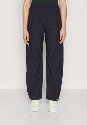 Person wearing loose black pants with elastic waistband and snap button cuffs, paired with white sneakers with yellow laces.