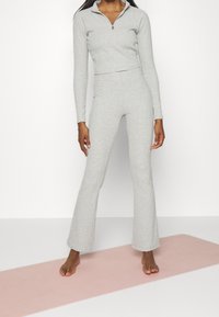 Donna in piedi che indossa un top zip-up a maniche lunghe grigio chiaro e pantaloni a zampa coordinati su un tappetino da yoga rosa, con sfondo bianco.