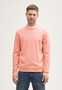 Helles korallenfarbenes Rundhals-Sweatshirt aus weichem Stoff, mit langen Ärmeln und gerippten Bündchen. Über einem weißen T-Shirt getragen, kombiniert mit Jeans.