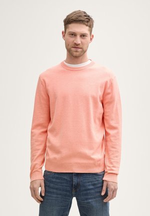 Un pull col rond corail clair en tissu doux, avec des manches longues et des poignets côtelés. Porté sur un t-shirt blanc, associé à un jean.