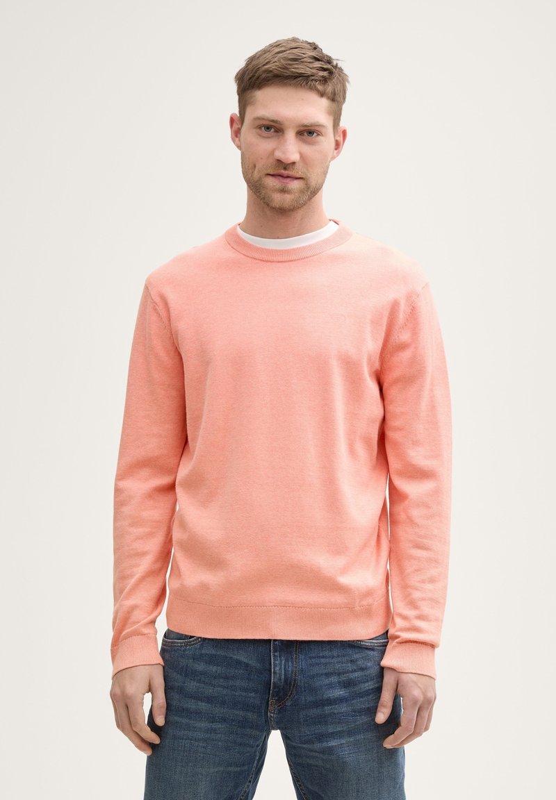 Helles korallenfarbenes Rundhals-Sweatshirt aus weichem Stoff, mit langen Ärmeln und gerippten Bündchen. Über einem weißen T-Shirt getragen, kombiniert mit Jeans.