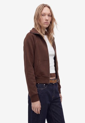 Ung kvinde med blondt hår iført en brun lynlåstrøje over en hvid skjorte og mørkeblå jeans, stående mod en ensfarvet baggrund.