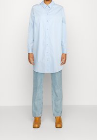 Person som bär en lång ljusblå knappad skjorta, ljusblå raka jeans och tanfärgade högklackade ankelstövlar mot en enkel bakgrund.