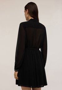Schwarzes Kleid mit einem plissierten Rock, langen durchsichtigen Ärmeln und einem taillierten Bund. Hervorzuhebende Details sind der Kragen und metallische Akzente.