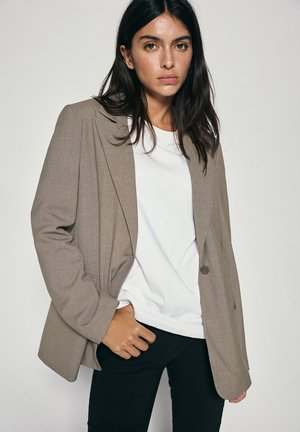 Femme aux longs cheveux foncés portant un blazer beige sur une chemise blanche et un pantalon noir, debout devant un fond clair uni.