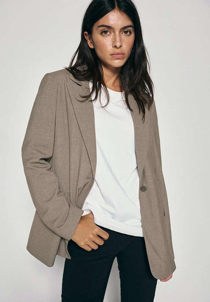 Femme aux longs cheveux foncés portant un blazer beige sur une chemise blanche et un pantalon noir, debout devant un fond clair uni.