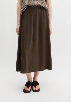 Kobieta ubrana w brązową spódnicę midi, dziergany top bez rękawów i ciemnobrązowe sandały na jednolitym jasnym tle.