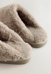 Chaussons en beige clair et duveteux avec une texture douce, en forme de orteil rond, et une base plate gris clair ; marqués "NEXT" sur le côté.