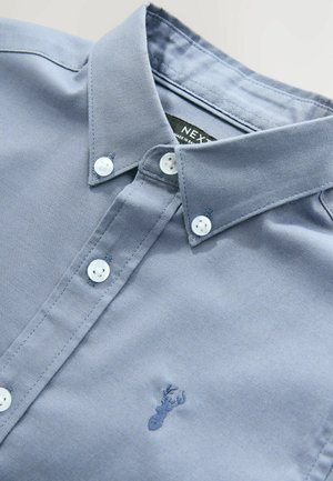 Chemise boutonnée bleu clair en tissu de coton. Présente des boutons blancs, un col et une petite broderie de homard bleu près de l'ourlet.