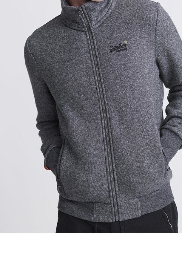 Homme portant une veste grise zippée avec les mains dans les poches, montrant un petit logo brodé sur la partie gauche de la poitrine.
