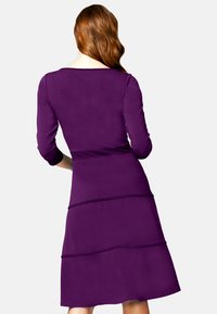 Violetti mekko pitkähihaisilla, istuvalla yläosalla ja levenevällä hameella, jossa on kerrokselliset kerrokset. Sileä kangas, jossa on kevyt kiilto ja pyöreä kaula-aukko.