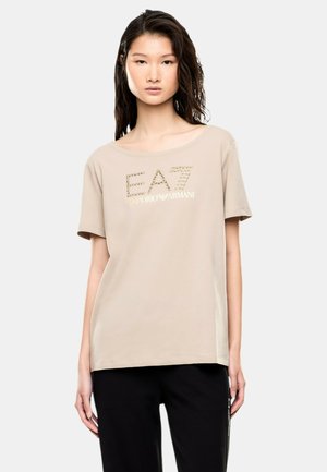 Donna con capelli lunghi e scuri indossa una t-shirt beige EA7 Emporio Armani e pantaloni neri, in piedi davanti a uno sfondo chiaro e uniforme.