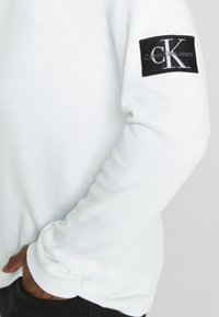 Bijela teksturirana majica s dugim rukavima, s crnim pravokutnim pečatom na lijevom rukavu s logotipom "CK Calvin Klein Jeans".