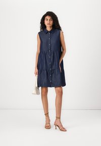 Denim dress in dark blue, sleeveless with a button-down front, tiered design, and collar. Model holding a light handbag.
