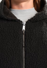 Musta fleece-takitakki, jossa on teksturoitu pinta, kontrastina sileä kangaskaulus ja metallinen vetoketju edessä.