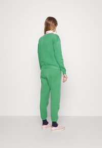 Zelená, strukturovaná mikina a odpovídající tepláky, s žebrovanými manžetami a pasem. Nosí se s růžovými teniskami a tmavými ponožkami.