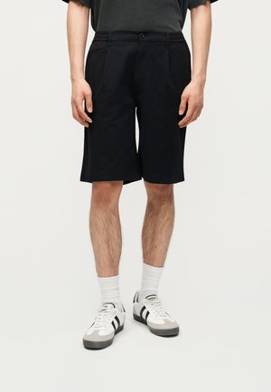 Osoba ubrana w czarne szorty do kolan, białe skarpetki i białe trampki z czarnymi paskami stoi na tle jednolitego tła.