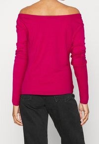 Haut fuchsia à manches longues et épaules dénudées, à la coupe ajustée et à la texture douce, associé à un jean foncé visible à la taille.