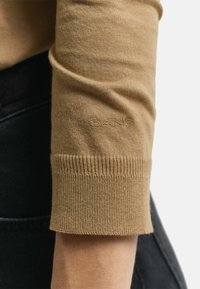 Manche de pull en tricot marron clair avec poignet côtelé, présentant un logo brodé ton sur ton. Texture lisse sur fond de tissu noir.