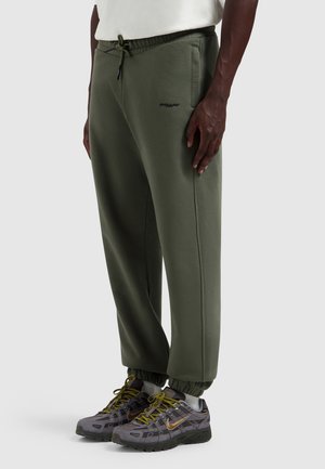 Man draagt olijfgroene joggerbroek met elastische manchetten en grijze Nike-sneakers met gele veters, staand op een effen achtergrond.