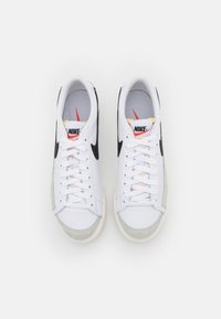 Paire de baskets blanches Nike avec un swoosh noir, bouts en suède gris et logo rouge sur la languette, vue de dessus sur fond blanc.
