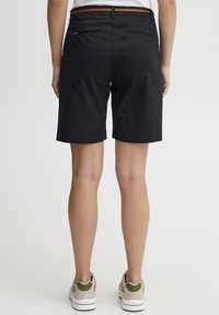 Shorts noirs en tissu léger, dotés d'une ceinture marron, de deux poches arrière et d'une coupe à mi-genoux. Portés avec des baskets décontractées.
