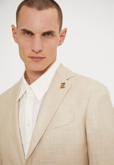 Blazer beige e texturizzato con motivo sottile, rever a punta singola e spilla color oro, indossato sopra una camicia bianca con bottoni.