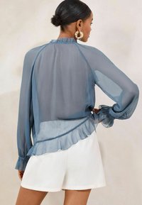 Sheer blue blouse with a ruffled hem, long sleeves, and elastic cuffs. Features a high neckline and a smooth, lightweight texture.