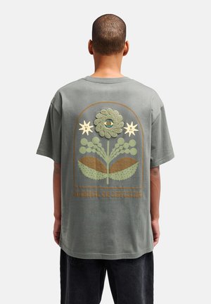 Personne portant un t-shirt gris avec un motif floral vert et marron et le texte « Balance of Opposites » au dos, vue de dos.