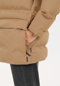 Hånd i lommen på en tan farvet pufferjakke med sorte lynlåstags, båret med mørkegrå bukser mod en simpel lys baggrund.
