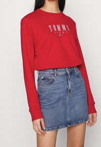 Červená košile s dlouhým rukávem a bílým logem "TOMMY JEANS", spárovaná s modrou denimovou sukní. Sukně má klasický střih a viditelné švy.