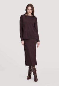 Top a maglia marrone scuro con scollatura a barca e spalle cadenti, abbinato a una gonna lunga aderente con spacco laterale.