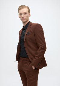 Bruin pak met een getailleerde pasvorm, bijpassende broek en een zwart shirt. Het pak heeft een enkele knoopsluiting en een pochet. Soepele textuur.