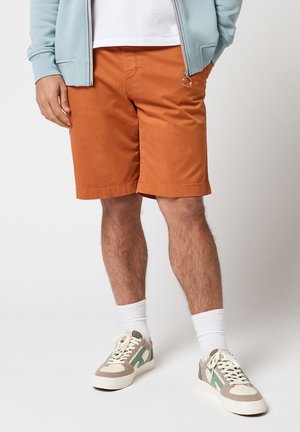 Homme portant un short couleur rouille, des chaussettes blanches, des baskets beige et vertes, et une veste zippée bleu clair, les mains dans les poches.