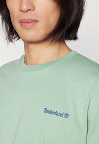 Lysegrønn crew-neck t-skjorte laget av mykt stoff, med en blå Timberland-logo på venstre brystområde. Glatt tekstur med korte ermer.
