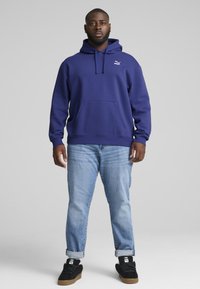 Felpa viola con tasca a marsupio e cappuccio regolabile, abbinata a jeans azzurri chiari e sneakers nere. Logo visibile sul petto.