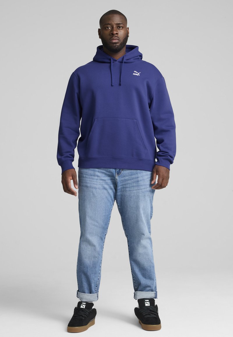 Felpa viola con tasca a marsupio e cappuccio regolabile, abbinata a jeans azzurri chiari e sneakers nere. Logo visibile sul petto.