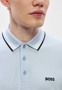 Ανοιχτό μπλε πόλο πουκάμισο με υφασμάτινη υφή, με ριγωτό γιακά σε μαύρο και άσπρο, τρία κουμπιά και μαύρο λογότυπο "BOSS" στο στήθος.