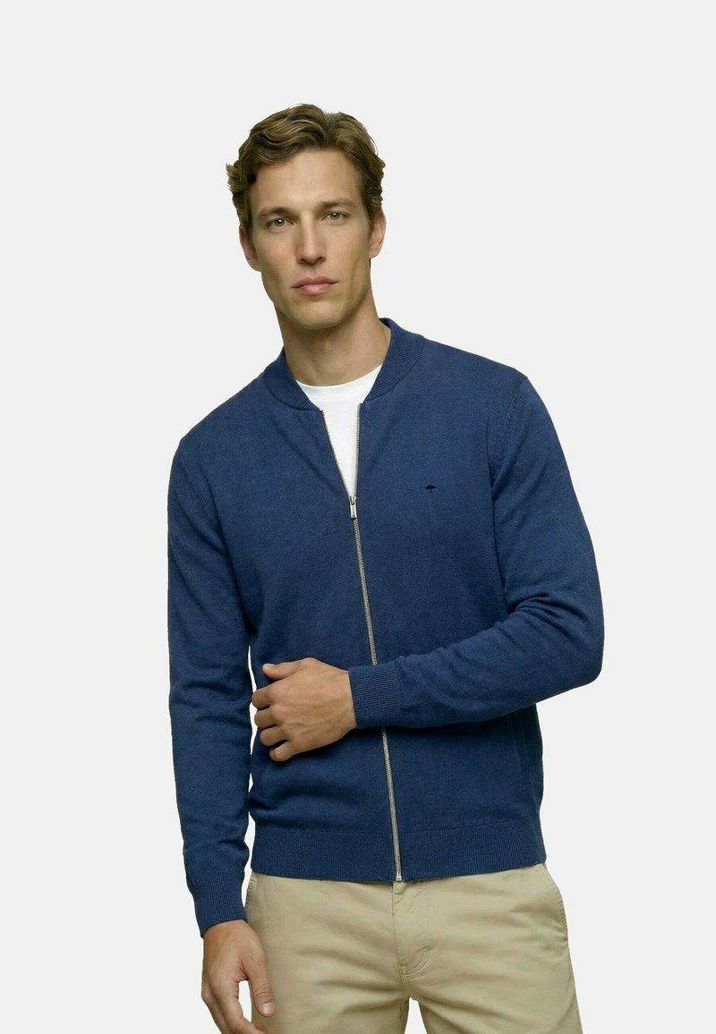 Mężczyzna w niebieskim kardiganie na zamek błyskawiczny, nałożonym na białą koszulę i beżowe spodnie, stojący z jedną ręką dotykającą drugiego ramienia.