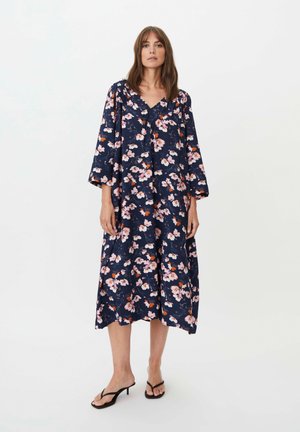 Kvinde iført en marineblå midikjole med pink og orange blomsterprint og løstsiddende lange ærmer, iført sorte sandaler med hæl.